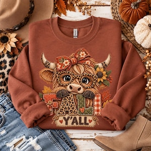 Op de afbeelding: Een roestkleurige sweater met ronde hals en een afbeelding van een Schotse hooglander met een bril met luipaardprint en een bloemenstrik. De koe staat boven de tekst "FALL Y'ALL" met letters in verschillende patronen zoals ruitjes en luipaard.