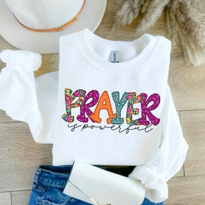 May include: White sweatshirt with the words "PRAYER is powerful" printed on the front. The word "PRAYER" is in colorful patchwork letters with floral designs.