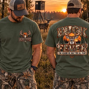May include: An olive green t-shirt featuring a small graphic on the front chest and a large graphic on the back. Both graphics display a deer head with antlers, camouflage lettering, and the text BUCK FEVER THE SEASON WE WAIT FOR ALL YEAR.
