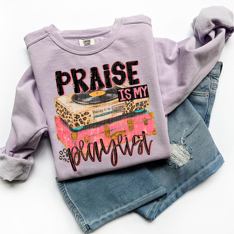 May include: A lilac Comfort Colors sweatshirt with the text "Praise is my playlist" printed on the front. The graphic features a vintage record player and stacked suitcases in leopard print and pink. Paired with denim shorts.