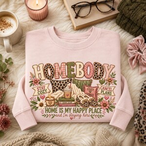 May include: A light pink crewneck sweatshirt featuring a graphic print with the text "HOMEBODY" in large letters, "STAY COZY", "CANCEL PLANS", and "HOME IS MY HAPPY PLACE AND I'M STAYING HERE". The design includes illustrations of pillows, books, and slippers.