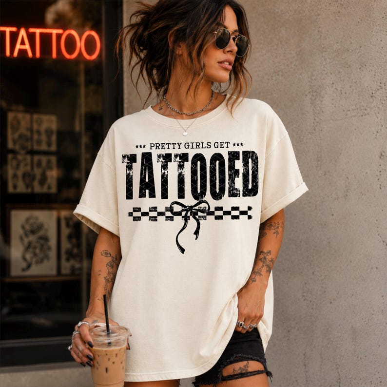 May include: An off-white short-sleeve t-shirt featuring the black text "PRETTY GIRLS GET TATTOOED" with a graphic of a bow and a checkered line. The shirt has a crew neckline and a relaxed fit, showcasing a bold, distressed-style typography design.