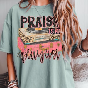 May include: A light teal t-shirt featuring the text "PRAISE IS MY PLAYLIST" in a layered design. The design includes a record player on top of a pink suitcase. The record player has a leopard print design.