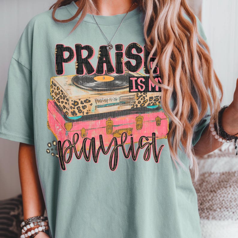 May include: A light teal t-shirt featuring the text "PRAISE IS MY PLAYLIST" in a layered design. The design includes a record player on top of a pink suitcase. The record player has a leopard print design.