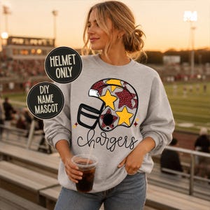 May include: A light gray crewneck sweatshirt featuring a graphic of a football helmet decorated with yellow and maroon stars. The word chargers is printed in black cursive script below the helmet. Two circular black labels read HELMET ONLY and DIY NAME MASCOT.