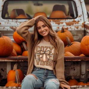 May include: A woman wearing a tan cardigan and a cream t-shirt with a brown and white plaid pumpkin graphic and the text "Cozy Vibes". She is sitting in the back of a truck bed surrounded by pumpkins.