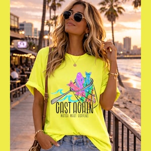 May include: A neon yellow t-shirt featuring a graphic of colorful fish in a net with the text "CAST AGAIN WATCH WHAT HAPPENS". The shirt has short sleeves and a relaxed fit. The background shows a coastal scene with palm trees and a sunset.