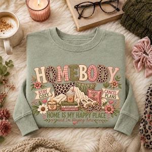 May include: A sage green crewneck sweatshirt featuring a graphic print with the text "HOMEBODY" and "HOME IS MY HAPPY PLACE and I'm staying here". The design includes illustrations of a leopard print pillow, a coffee cup, books, and slippers.