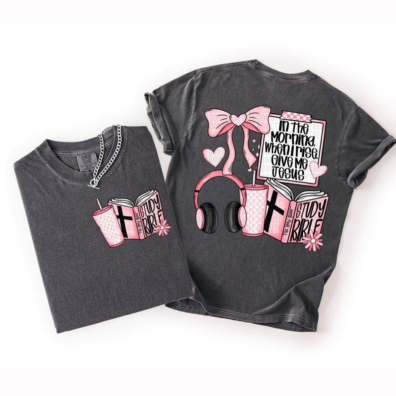 May include: Two charcoal gray t-shirts with pink and white graphics. One shirt features a book, drink, and flower. The other has a bow, headphones, drink, and the text "In the morning when I rise, give me Jesus."