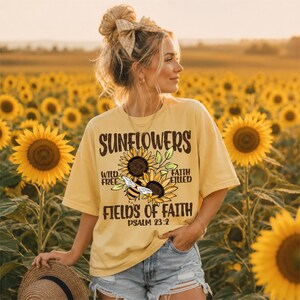 Op de afbeelding: Een lichtgeel T-shirt met korte mouwen en een afbeelding van twee zonnebloemen, een bij en de tekst: SUNFLOWERS, WILD FREE, FAITH FILLED, FIELDS OF FAITH, PSALM 23:2. Het T-shirt wordt gedragen met een lichtblauwe spijkerbroek met rafelige randen.