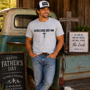 May include: A man wearing a gray t-shirt with the text "FAITHFUL FATHER. GODLY MAN." and a black cross. He is also wearing a black and white hat with the word "FREEDOM". A sign reads "HAPPY FATHER'S DAY".