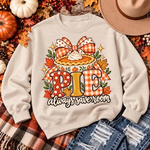 Puede incluir: Una sudadera beige de cuello redondo con un gráfico de tarta de calabaza con nata montada, un gran lazo a cuadros naranja y blanco, hojas de otoño y el texto PIE always save room en patrones de color naranja, rojo y amarillo.