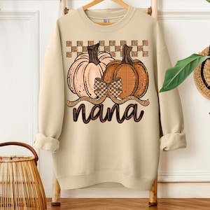 May include: Beige sweatshirt with a pumpkin design, bow, and the word "nana". The pumpkins are orange and peach, with a checkered pattern above. The sweatshirt has long sleeves and a crew neck. A cozy autumn themed top.