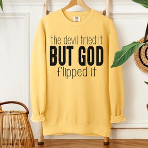 May include: A yellow sweatshirt with the text "the devil tried it BUT GOD flipped it" in black. The sweatshirt is hanging on a wooden hanger against a neutral background. The sleeves are cuffed.