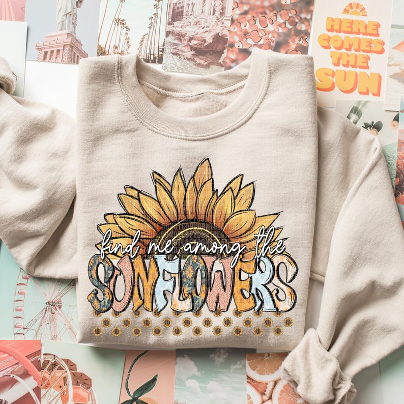 May include: A beige crewneck sweatshirt featuring a large sunflower graphic with the text "find me among the sunflowers". The sunflower is yellow, brown, and black, with the text in various colors. The sweatshirt is folded.