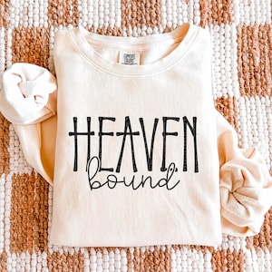 May include: Cream-coloured sweatshirt with the words "HEAVEN bound" in black script. The sweatshirt has long sleeves and a crew neck. The brand "Comfort Colors" is visible on the tag. The background is a woven pattern.