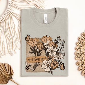 May include: A light gray t-shirt featuring a floral design with butterflies and a bee. The design includes flowers in shades of brown and white, with the words "good things grow" on a banner.