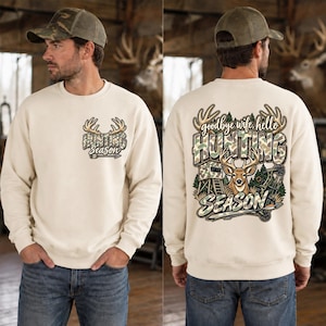 Könnte beinhalten: Ein cremefarbenes Sweatshirt mit Rundhalsausschnitt und einer kleinen Grafik auf der linken Brust mit Hirschgeweih und dem Text "HUNTING SEASON". Die Rückseite zeigt eine große Grafik mit Geweih, einem Hirsch, einem Hochsitz und "goodbye wife, hello HUNTING SEASON".