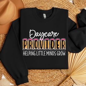 May include: Black sweatshirt with the words "Daycare PROVIDER" in white and yellow, with pencil graphics. The text "HELPING LITTLE MINDS GROW" is below the main text. The sweatshirt is displayed on a woven surface.