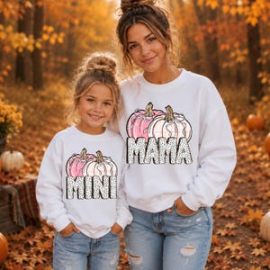Peut inclure: Deux sweat-shirts blancs à manches longues avec un motif de deux citrouilles ornées de nœuds et le texte MAMA sur l'un et MINI sur l'autre. Le texte présente un motif à pois noirs et blancs. Les citrouilles sont de couleur rose et blanche.