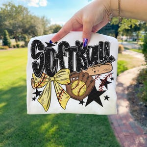 May include: White t-shirt featuring the word "SOFTBALL" in a black and white design. The shirt also includes a yellow bow, a baseball glove, a baseball, a baseball bat, and black stars. The design is likely for a softball player or fan.