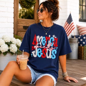 K&ouml;nnte beinhalten: Marineblaues T-Shirt mit dem Text "America needs Jesus" in Rot, Wei&szlig; und Blau. Das Design beinhaltet Sterne und Kreuze. Das Shirt wird mit Jeansshorts getragen. Im Hintergrund ist eine amerikanische Flagge zu sehen.