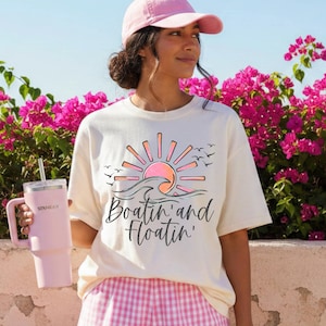 May include: Cream-colored t-shirt with a graphic of a sunset over ocean waves, the text "Boatin' and Floatin'", and flying birds. A pink tumbler with a straw is held in the image. The person is wearing a pink baseball cap and pink and white checkered shorts.