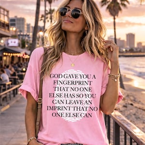 Può includere: T-shirt rosa chiaro con il testo bianco "GOD GAVE YOU A FINGERPRINT THAT NO ONE ELSE HAS SO YOU CAN LEAVE AN IMPRINT THAT NO ONE ELSE CAN". La maglietta ha una grafica di impronta digitale bianca.