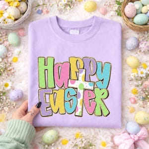 May include: A light purple sweatshirt with the words "Happy Easter" in colorful, bubble letters. The letters are outlined in gold and feature a cross with pastel polka dots. The sweatshirt is surrounded by Easter eggs and flowers.