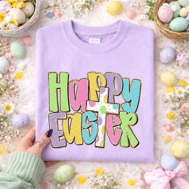 May include: A light purple sweatshirt with the words "Happy Easter" in colorful, bubble letters. The letters are outlined in gold and feature a cross with pastel polka dots. The sweatshirt is surrounded by Easter eggs and flowers.