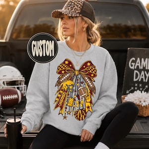 May include: A light gray crewneck sweatshirt featuring a large graphic of a decorative bow adorned with football-themed elements including a helmet, a football, a whistle, a pom-pom, and the word CHARGERS. A circular badge with the text CUSTOM is visible.
