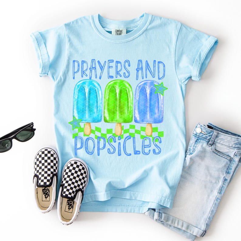 Puede incluir: Camiseta azul claro con las palabras "Prayers and Popsicles" y un gr&aacute;fico de tres polos de colores. La imagen incluye pantalones cortos de mezclilla, zapatos sin cordones a cuadros y gafas de sol.