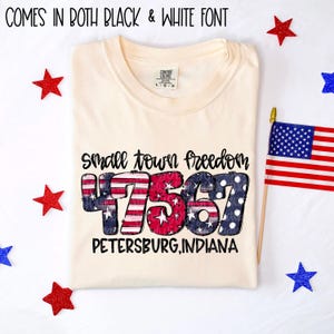 Può includere: T-shirt color crema con la scritta "Small Town Freedom 47567 Petersburg, Indiana". I numeri sono disegnati con un tema patriottico rosso, bianco e blu. La maglietta è circondata da stelle rosse e blu e una piccola bandiera americana.