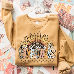 May include: Mustard yellow sweatshirt featuring a large sunflower graphic with the text "find me among the SUNFLOWERS". The design includes a detailed sunflower with yellow petals and a brown center. The text is in a mix of colors and fonts.