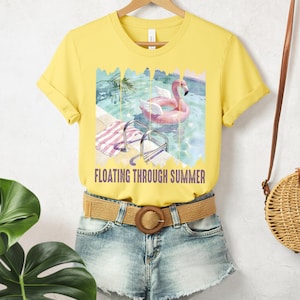 Może przedstawiać: Żółty t-shirt z grafiką różowego flaminga w basenie, z napisem "FLOATING THROUGH SUMMER". Koszulka jest połączona z dżinsowymi szortami i plecionym paskiem z okrągłą klamrą. W tle wisi słomiana torba.