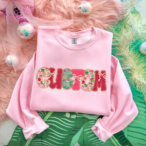 May include: A light pink sweatshirt with the word "CUTIE" embroidered across the front in a festive design. The letters are red, green, and pink with gold accents. The sweatshirt is displayed on a pink fluffy surface with Christmas ornaments.