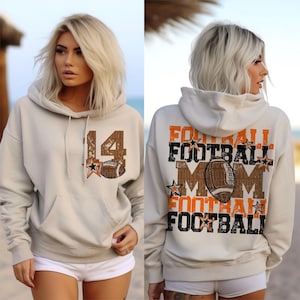 May include: A light beige hoodie with a front pocket and drawstring. The front features the number 14, a star, and a football graphic. The back of the hoodie displays the words "FOOTBALL MOM" and a football graphic in orange and brown.