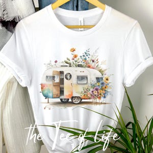 Op de afbeelding: Wit T-shirt met een aquarel-illustratie van een vintage camper met bloemen die erop groeien. De tekst "the tee of life" is onder de afbeelding gedrukt.