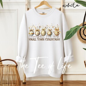 May include: White crewneck sweatshirt featuring a festive design. The design includes gold and white Christmas ornaments with bows, and the text "Farmingville, New York SMALL TOWN CHRISTMAS". The words "the Tee of Life" are also visible.