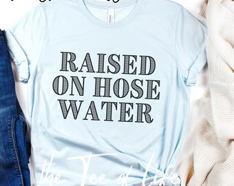 May include: A light blue t-shirt with the text "RAISED ON HOSE WATER" printed in black. The shirt is made of a soft, comfortable material.