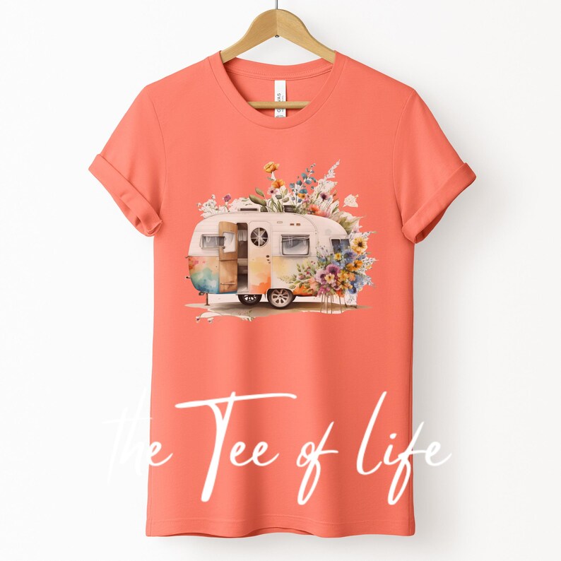 Op de afbeelding: Een koraalkleurig T-shirt met een aquarel-illustratie van een vintage camper die omringd is door kleurrijke bloemen. De tekst "The Tee of Life" is gedrukt op de onderkant van het shirt.