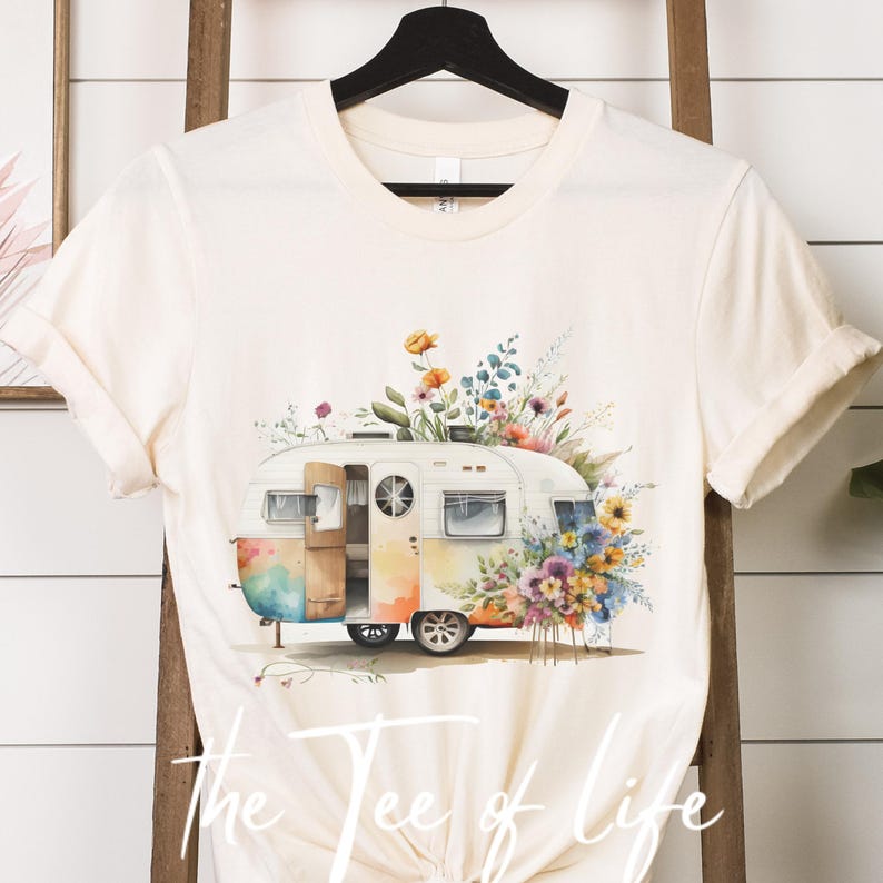 Op de afbeelding: Een witte vintage camper met een aquarel bloemenontwerp. De camper is omringd door kleurrijke bloemen en groen. De camper heeft een open deur en een zichtbaar raam. Het ontwerp is op een cr&egrave;mekleurig T-shirt.
