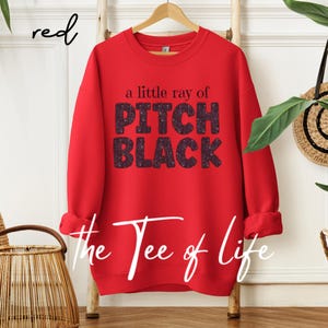 May include: A red sweatshirt with the text "a little ray of PITCH BLACK" in black glitter lettering. The words "the Tee of life" are written in white script at the bottom. The word "red" is written in red script in the upper left corner.