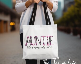 May include: White tote bag with black handles. The bag has a pink and black floral design that spells out "AUNTIE like a mom, only cooler".