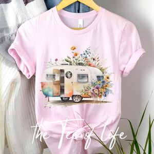 Op de afbeelding: Een roze T-shirt met een aquarel-illustratie van een vintage camper die omringd is door kleurrijke bloemen. De camper is wit met een regenboogkleurig ontwerp aan de zijkant. De tekst "The Test of Life" is gedrukt op de onderkant van het shirt.