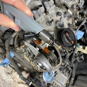 Puede incluir: Primer plano de un inyector de combustible gris que se sostiene con una mano. El inyector está conectado a un cable negro y tiene una punta de metal. El inyector se está instalando en un motor de automóvil.