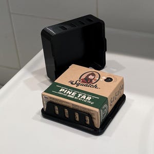 Puede incluir: Un jabonera negra con una barra de jabón marrón en su interior. El jabón está etiquetado como "The Squatch. Jabón natural para hombres/Savon Naturel Pour Hommes Pine Tar Peso neto 5 oz (141 g)"