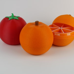 Könnte beinhalten: Drei fruchtförmige Behälter: eine rote Tomate mit grünem Stiel und zwei Orangen. Eine Orange ist geöffnet und zeigt ein segmentiertes Inneres mit Wochentagen. Die Behälter dienen wahrscheinlich zur Aufbewahrung kleiner Gegenstände.