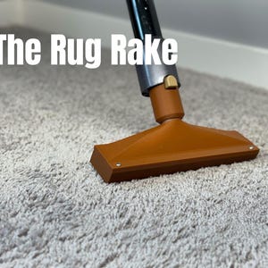 May include: An orange rug rake with a silver and black handle is shown on a gray carpet. The text "The Rug Rake" is displayed in white letters at the top of the image. The rake is designed for cleaning carpets.