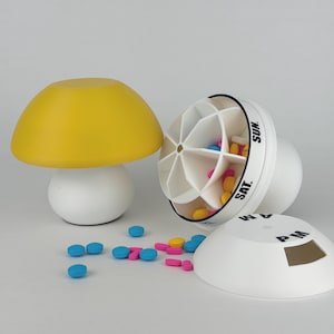 May include: A white and yellow mushroom-shaped pill organiser with a removable lid. The pill organiser is open, revealing compartments filled with colourful pills. The lid is labelled with "AM" and "PM".